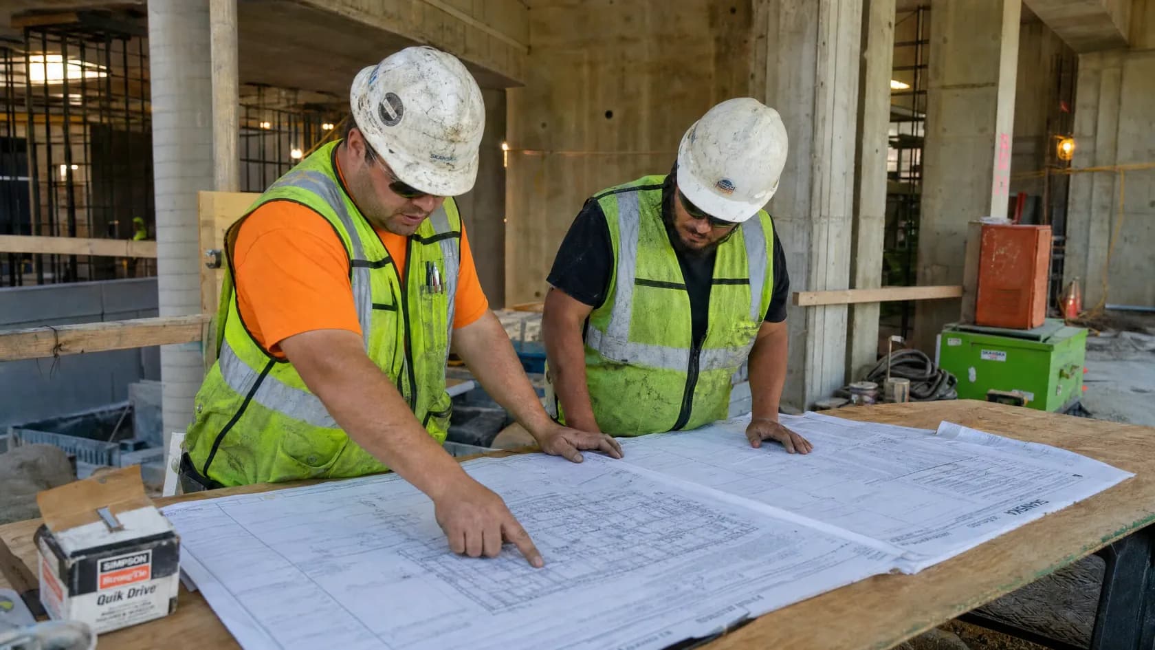 Two workers in hard hats reading a plan set spread across a plywood bench on an active jobsite